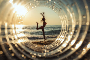 Girl doing a gymnastic pose on the beach at sunset, seen through a circular light effect