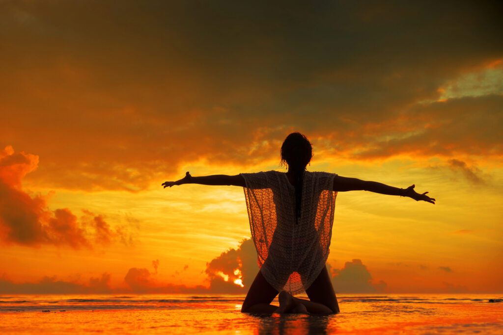 Woman kneeling on the beach with arms outstretched at sunset